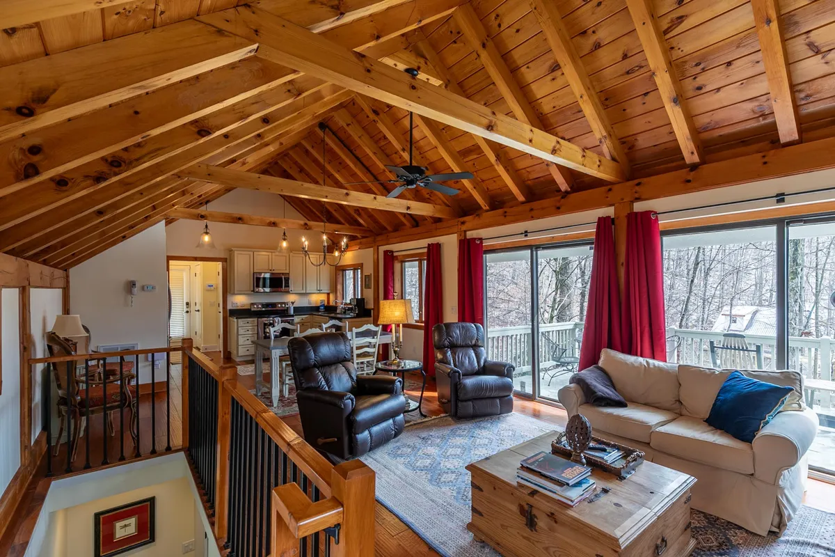 A sun-drenched great room featuring exposed wooden beams overhead, large windows framing views of the forest, and a comfortably arranged seating area gathered around a central coffee table. The inviting atmosphere encourages relaxation and conversation, with natural light bathing the rustic furnishings and the outdoors always in view.