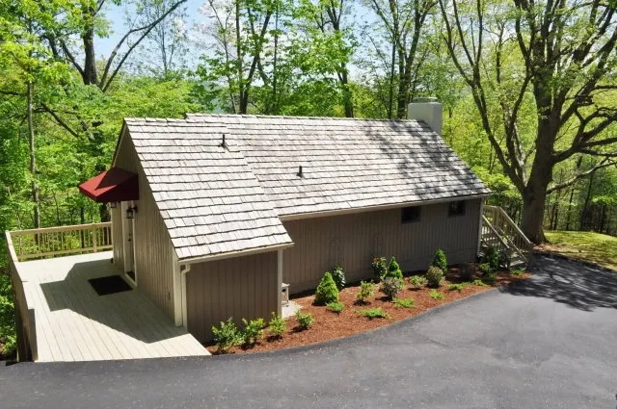 The image shows the exterior of a home with a welcoming deck and lush surrounding greenery. The deck is spacious, suitable for outdoor gatherings, barbecues, or simply relaxing in nature. The scene is peaceful, and the home appears to be part of a cluster, suggesting proximity to other cabins.