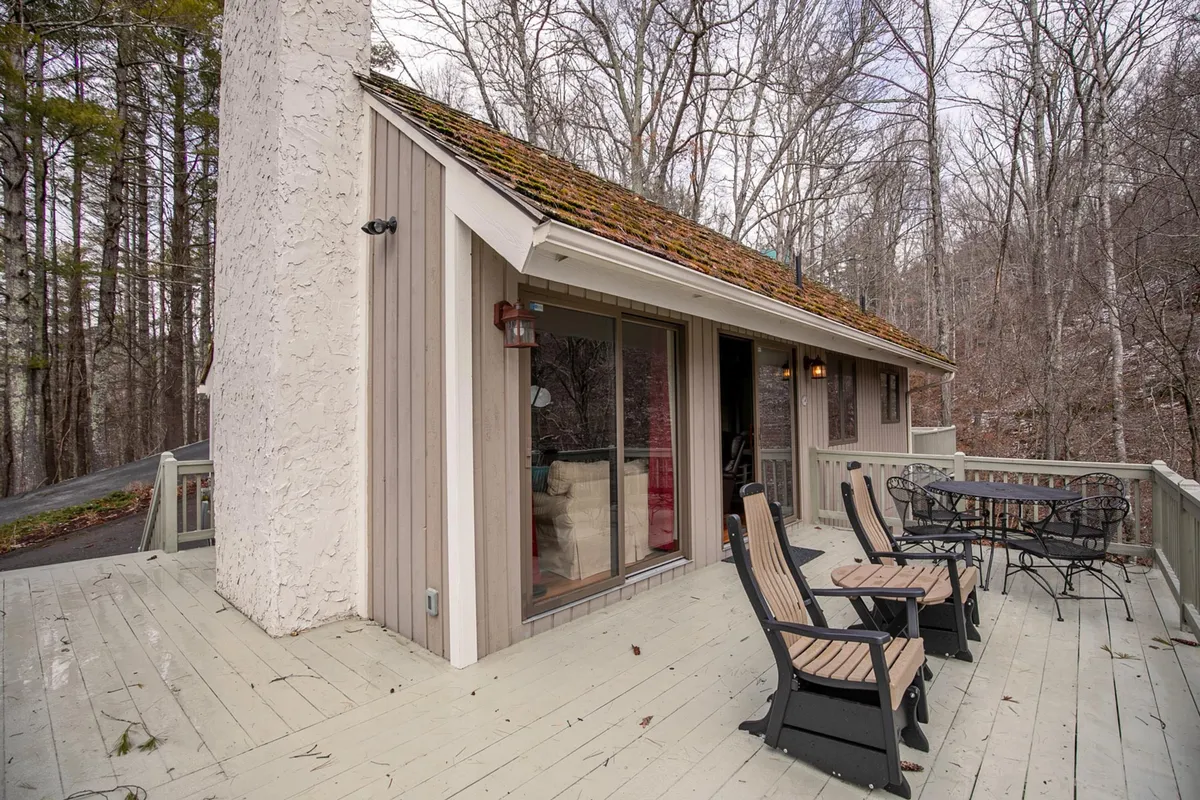 This image features the deck area of the house, framed by large windows and a visible chimney, all surrounded by mature trees. The deck appears spacious and is set up for relaxing and dining, capturing the essence of outdoor mountain living.