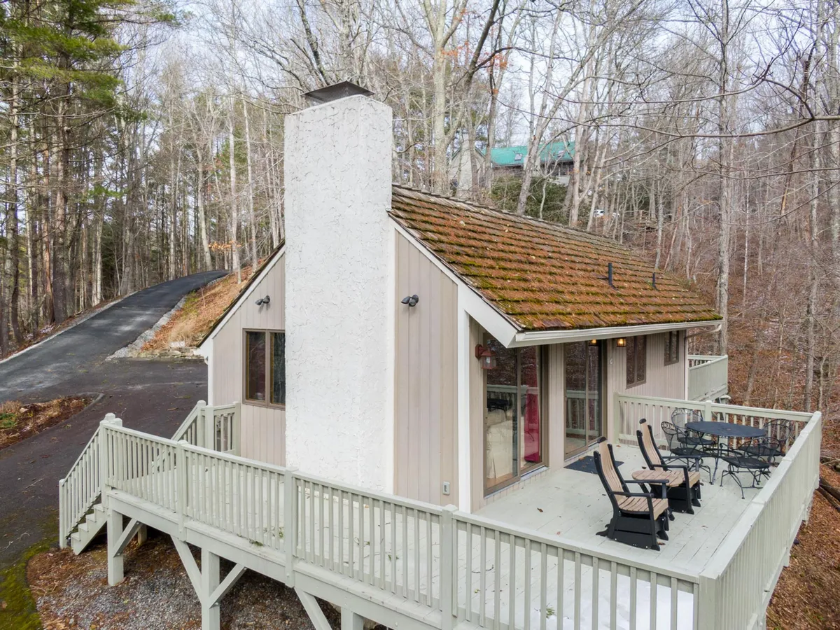 This image showcases the exterior of a cozy mountain cabin surrounded by trees, featuring a large chimney and a spacious deck with outdoor seating. The setting suggests a tranquil place suited for outdoor meals, gatherings, and relaxation.