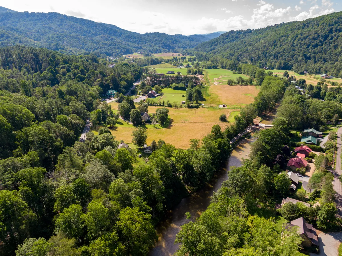 A panoramic vista of the High Country with rolling green fields, a meandering river, scattered houses, and majestic mountains stretching into the distance. The scene is bathed in serene, early morning light, and hints of village life are visible nearby.