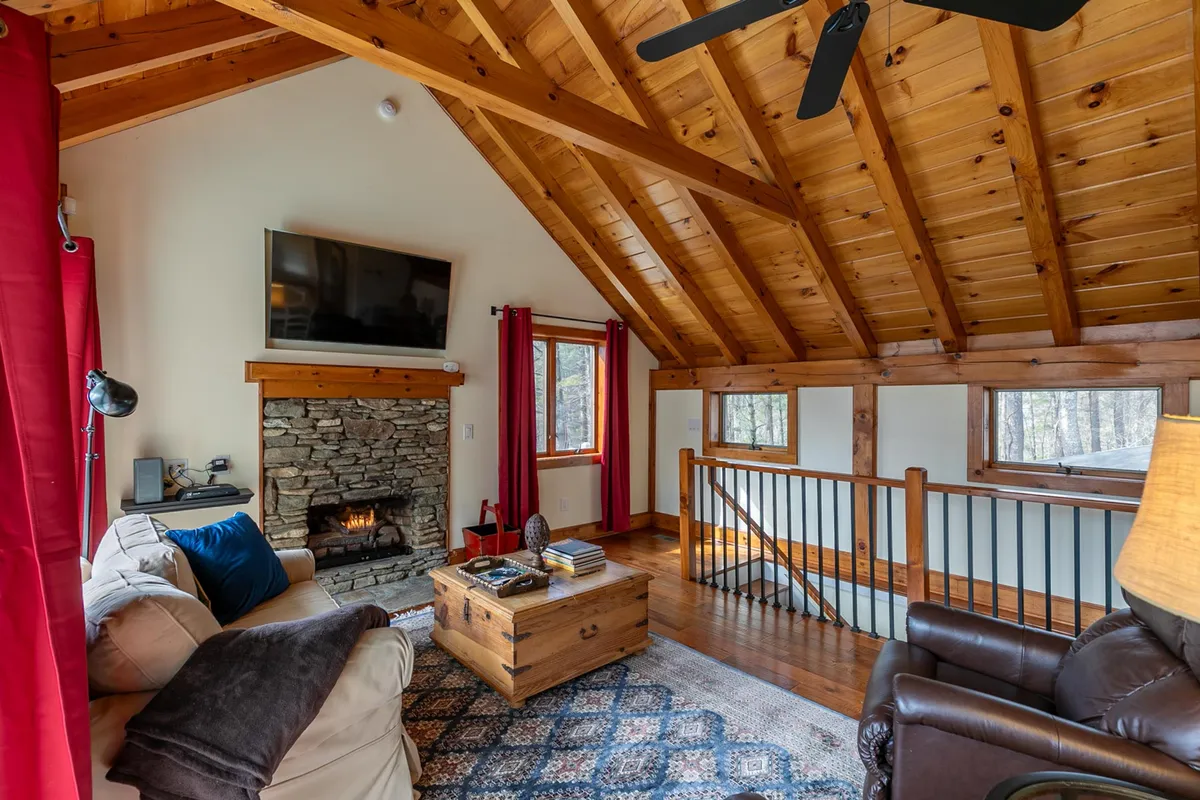 A cozy living area featuring a stone fireplace, wooden beams across the ceiling, plush seating, and expansive windows letting in natural light. The arrangement invites guests to settle in for warmth and intimate gatherings, with the fireplace as a focal point for cozy evenings or movie nights.