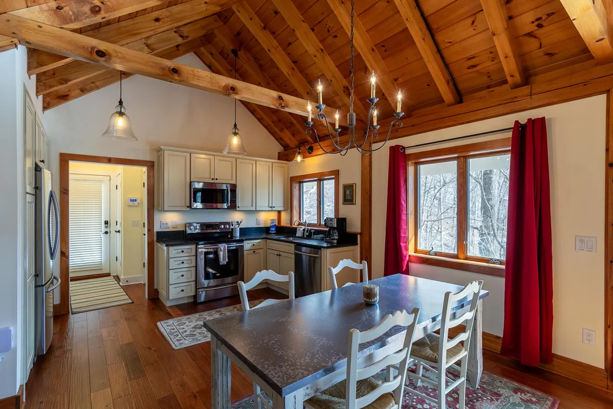 A welcoming dining area seamlessly connected to the kitchen, featuring a sturdy wooden dining table, kitchen appliances, a charming chandelier, large windows with mountain or forest views, and exposed wooden beams above. The space is designed for shared meals, conversation, and effortless flow between cooking and dining.