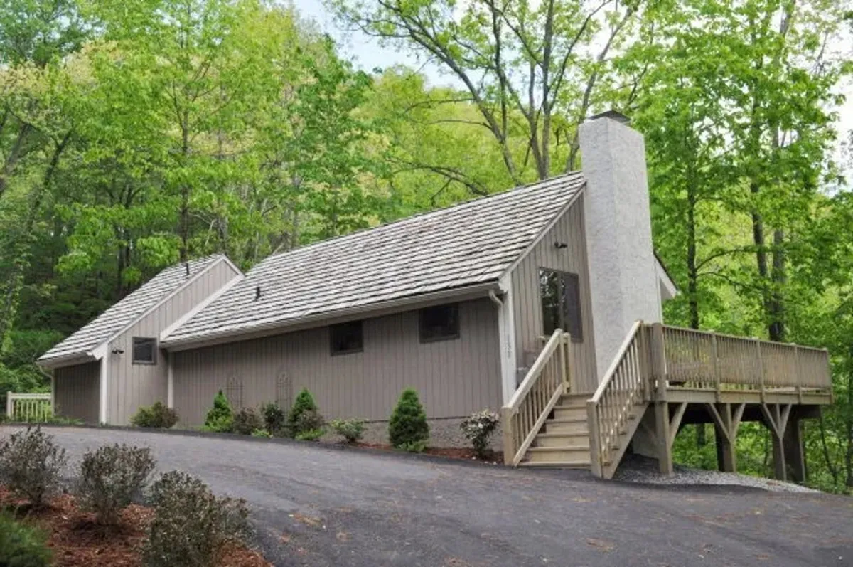 This exterior image features a wooden deck, a sloped roof, and abundant greenery around the cabin. The deck provides an inviting space for relaxing, enjoying nature, or hosting group activities, all set in a tranquil environment.